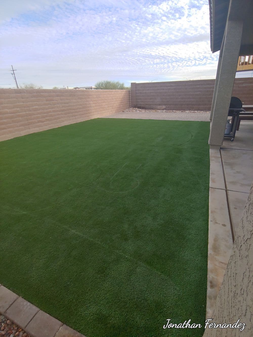 Lush green artificial turf lawn in a backyard with beige stone walls and clear sky.