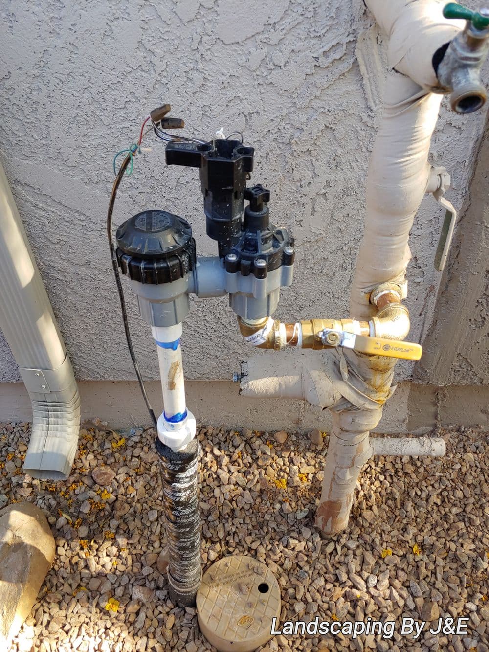 Irrigation valve system installed on exterior wall with plumbing connections and landscape gravel.