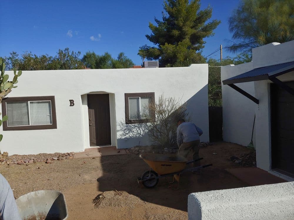 Person working with a wheelbarrow in a desert yard near a white adobe-style house.