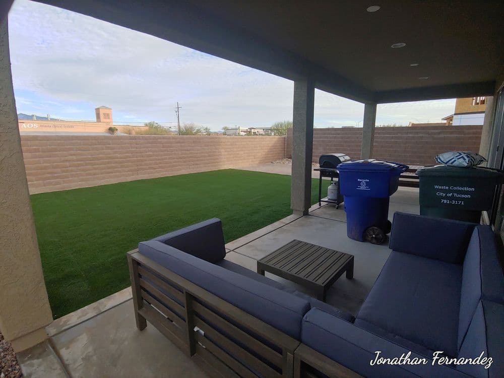 Patio with a sofa set, table, and trash bin, featuring artificial grass and a wall backdrop.