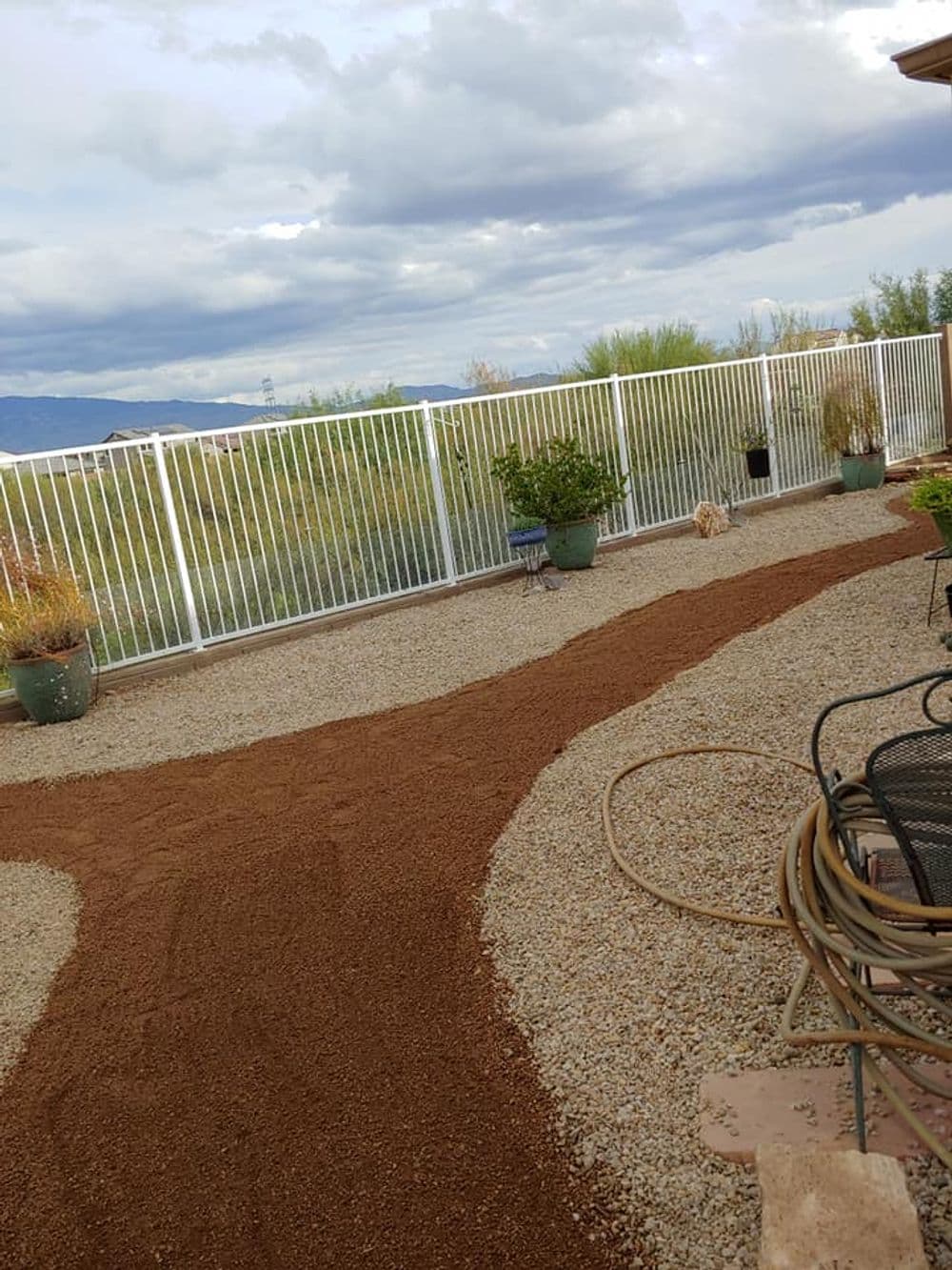 Desert landscape with gravel pathways, potted plants, and a white fence under cloudy skies.